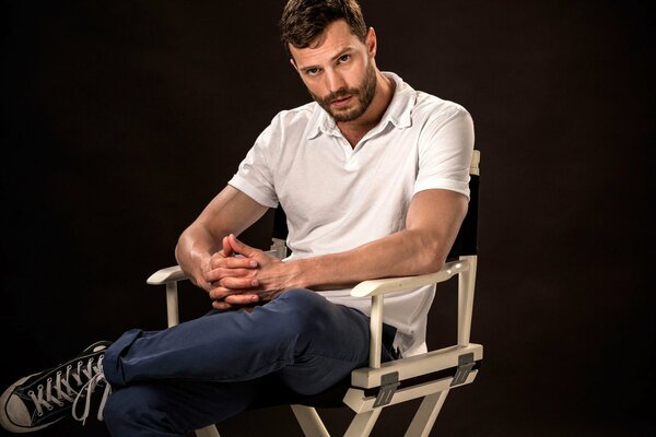 El actor, modelo y músico Jamie Dornan, con una camiseta blanca, pantalones azules y Zapatillas negras, se sienta en una silla de madera con los dedos cruzados y la pierna izquierda sobre la rodilla derecha sobre un fondo negro