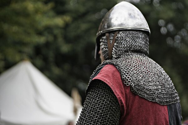 A warrior in helmet, armor and chain mail. There is a tent in the background