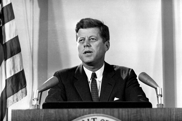 Kennedy en la tribuna con dos micrófonos y una bandera