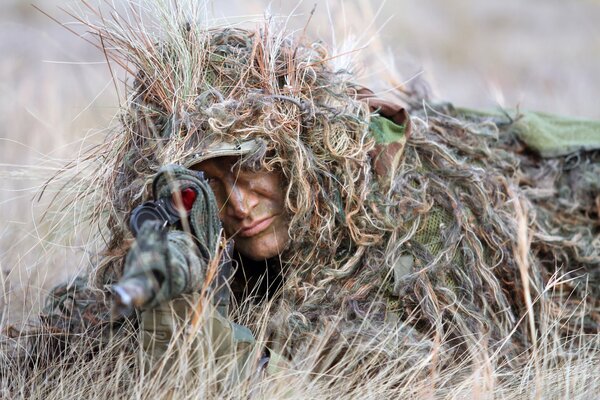 The sniper is disguised by grass and aims at the rifle