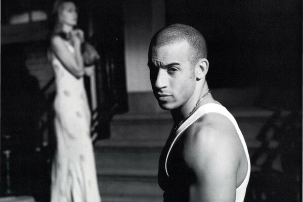 Vin Diesel steht im Profil und schaut in die Kamera im Hintergrund steht eine Frau