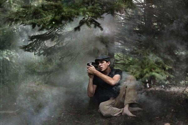 A young photographer is sitting barefoot on the ground in a foggy coniferous forest and taking pictures