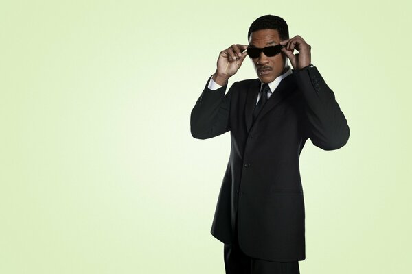 A dark-skinned man in black glasses and a suit poses mysteriously on a green background