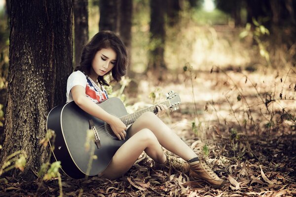 A girl is playing the guitar, leaning against a tree