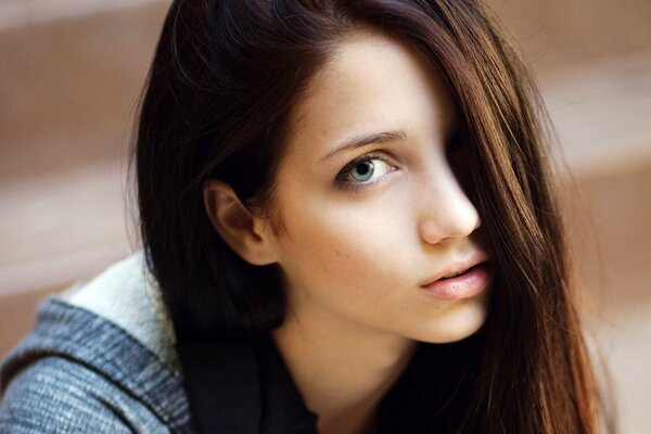 Beautiful blue-eyed brown-haired model Emily Rudd, covering her left eye with a strand of hair, looks sweetly into the frame