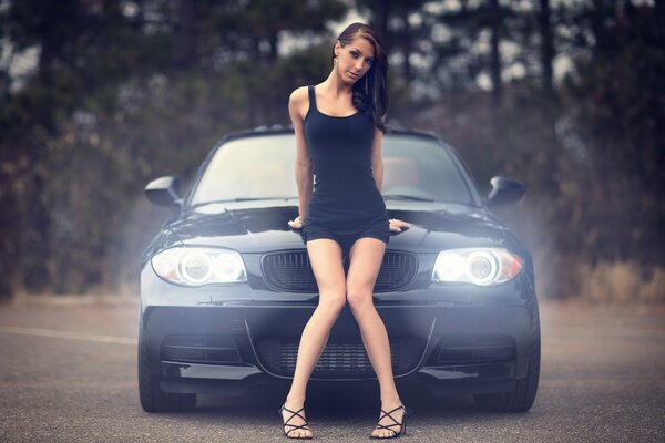 A brown-haired woman in a black tight dress sits on the hood of a BMW with her knees joined