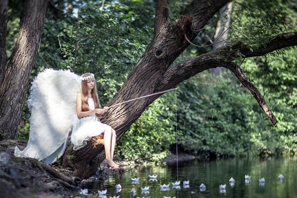 A girl in the image of an angel with white wings sitting on a tree with a fishing rod over a pond summer landscape