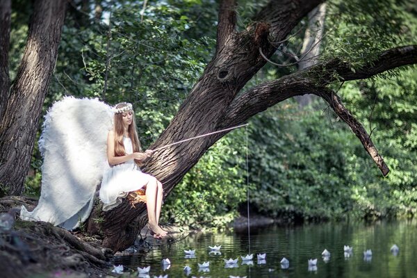 Una chica en la forma de un ángel con alas blancas sentado en un árbol con una caña de pescar sobre un estanque paisaje de verano