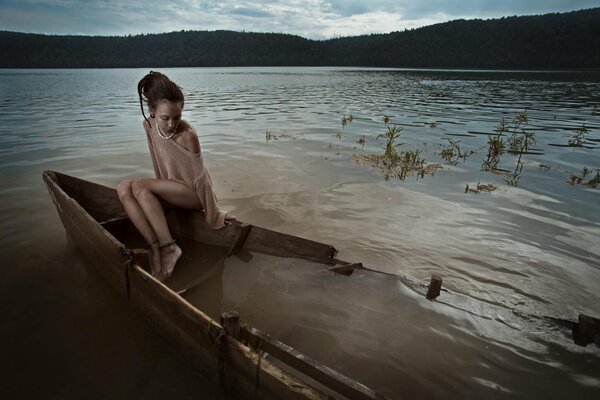 A lonely girl is sitting in a flooded boat on the shore of a lake