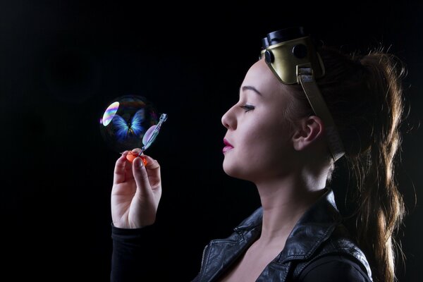 Une fille dans des lunettes de moto regarde une bulle de savon dans laquelle un papillon bleu est enfermé