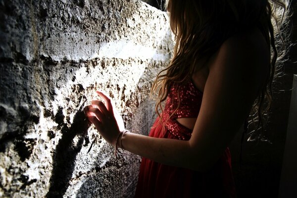 A brown-haired woman in a shiny red top stands near a concrete wall, on which a bright ray of light falls