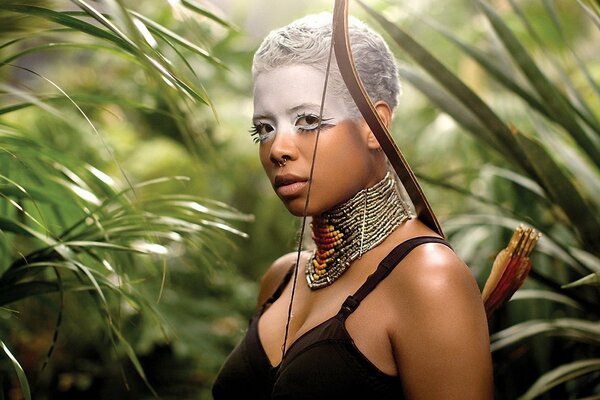 A dark-skinned woman with war paint on her head in an ethnic necklace and a war bow on her shoulder stands in the middle of the jungle