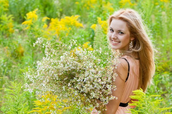 A smiling girl in long earrings stands with a bouquet of white wildflowers in a pink dress among the greenery