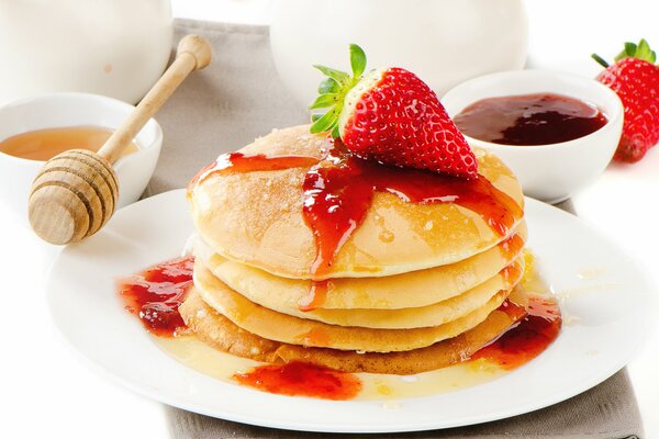 A mountain of pancakes on a plate with strawberry jam and honey