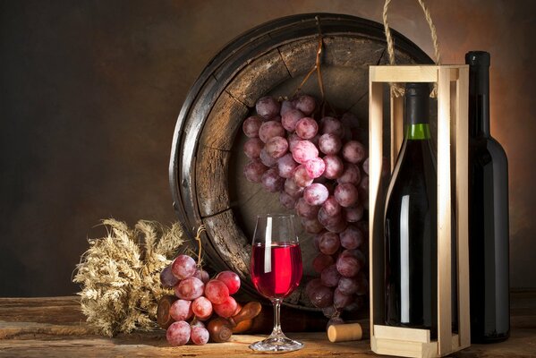 На столе бутылка в коробке и бокал вина, а рядом в бочонке гроздь винограда