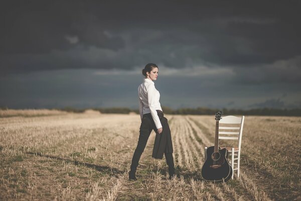 A girl in a business white shirt on the field with a guitar and a chair