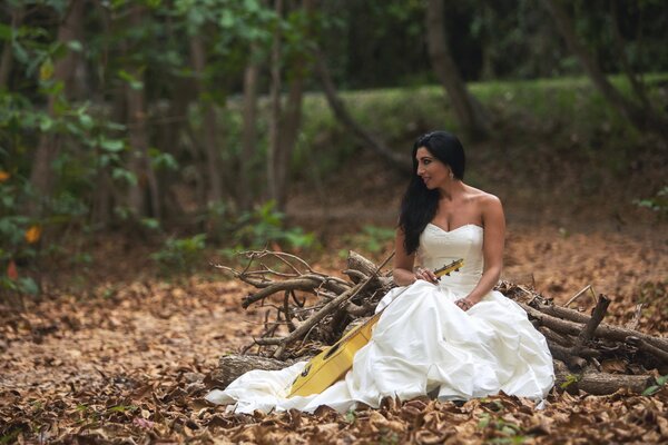 A girl in a wedding dress is sitting in the woods with a guitar