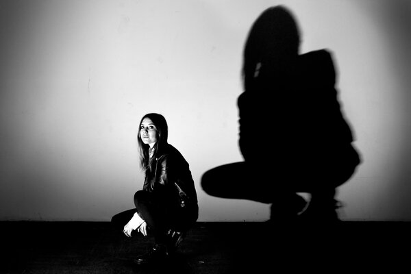 Девушка сидит на корточках на сером фоне отражается её чёрная тень