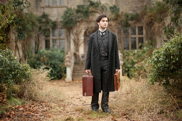 Daniel Radcliffe avec des valises en costume noir en face d un manoir abandonné