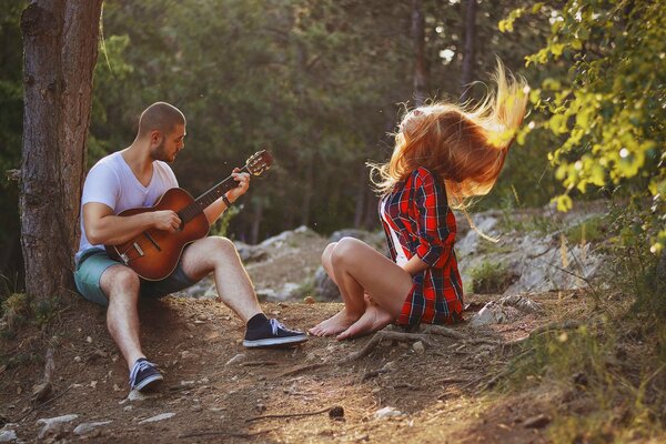 A guy plays guitar next to a girl in the woods