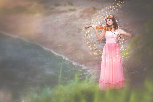 A girl in a pink dress plays a melody on a violin on the shore