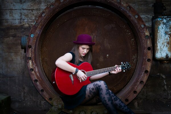 Una chica toca una guitarra roja, sentada en una rueda oxidada