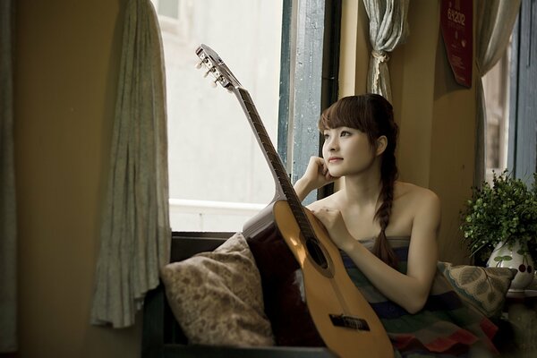 A girl is sitting with a guitar by the window