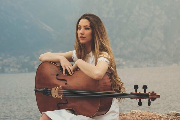 A girl sitting on the shore of a lake with a cello on her lap