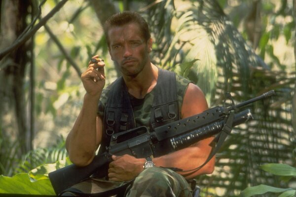 El actor, productor y director rold Schwarzenegger con un cigarro y una ametralladora en la mano