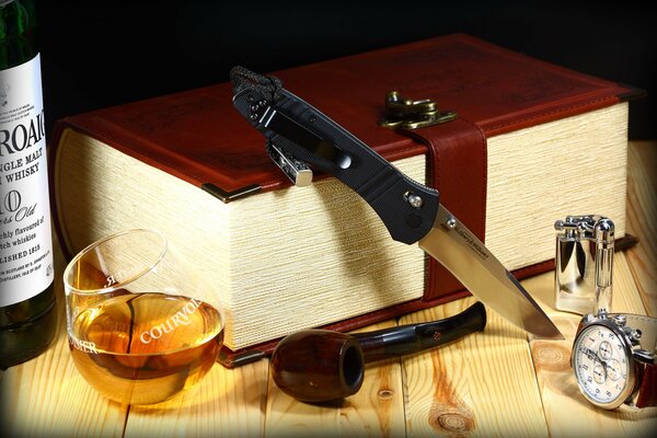 The knife is lying on a thick book and there is a whiskey with a pipe next to it