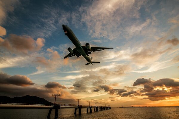 The plane is low over the river and the bridge at sunset