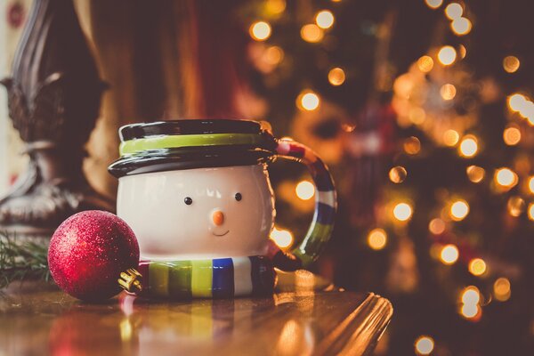 Becher- Schneemann und roter Christbaumkugel auf Kamode vor dem Hintergrund von Lichtern