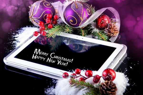 A white tablet with congratulations on the New Year and Christmas purple Christmas balls small piles of snow green Christmas tree branches cones and red berries on a dark background