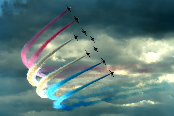 Plusieurs avions de combat, sur fond de nuages, mènent derrière eux des panaches multicolores