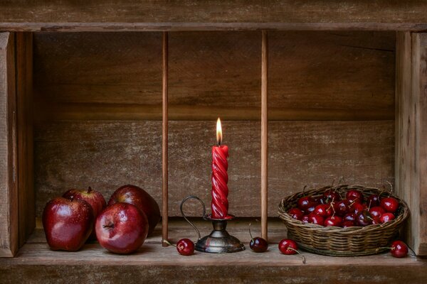 Una vela roja torcida se quema entre las cerezas en una canasta y las manzanas en un estante de madera