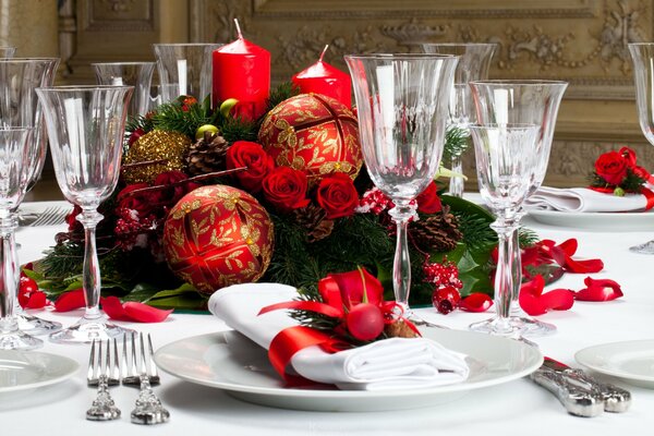 Tischdekoration des Neujahrs mit Forforgläsern mit weißen Servietten und einem mit leuchtend roten großen Kugeln geschmückten Christbaumkranz vor dem Hintergrund hoher roter Kerzen