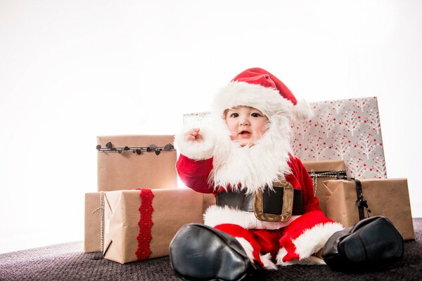 Un niño pequeño con un disfraz de papá Noel se sienta en el fondo de Koro ok con regalos y felicita a todos con un feliz año Nuevo y feliz Navidad