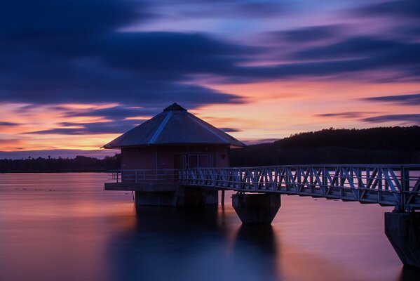 A peaceful picture. A bridge on the water with a house, against the background of the evening sky. In the distance you can see the forest