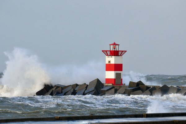 A sea wave breaks on a rocky base with a red and white lighthouse