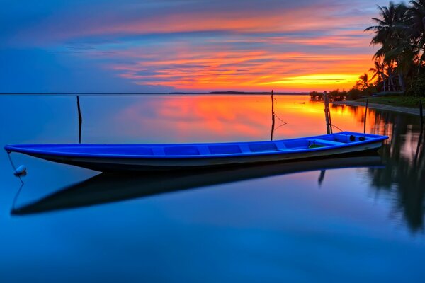 Barco largo en la tranquila superficie del agua en el fondo de palmeras y puesta de sol