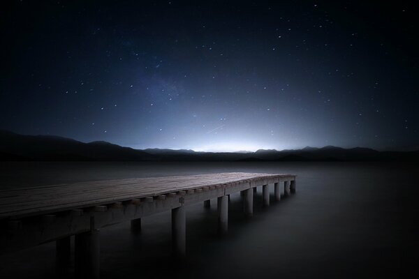 Sternenhimmel über dem Pier in der Nähe des Sees. In der Ferne sieht man Berge